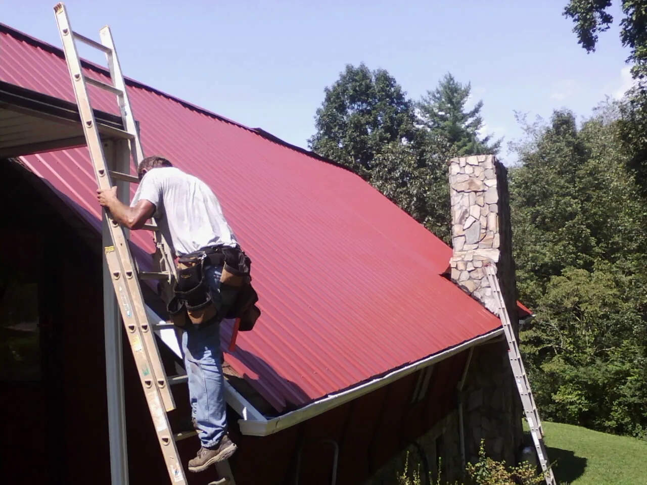 Roofer on a ladder beside a red metal roof and stone chimney — Soffit & Fascia Repair by Roofing Brothers in Mount Gilead